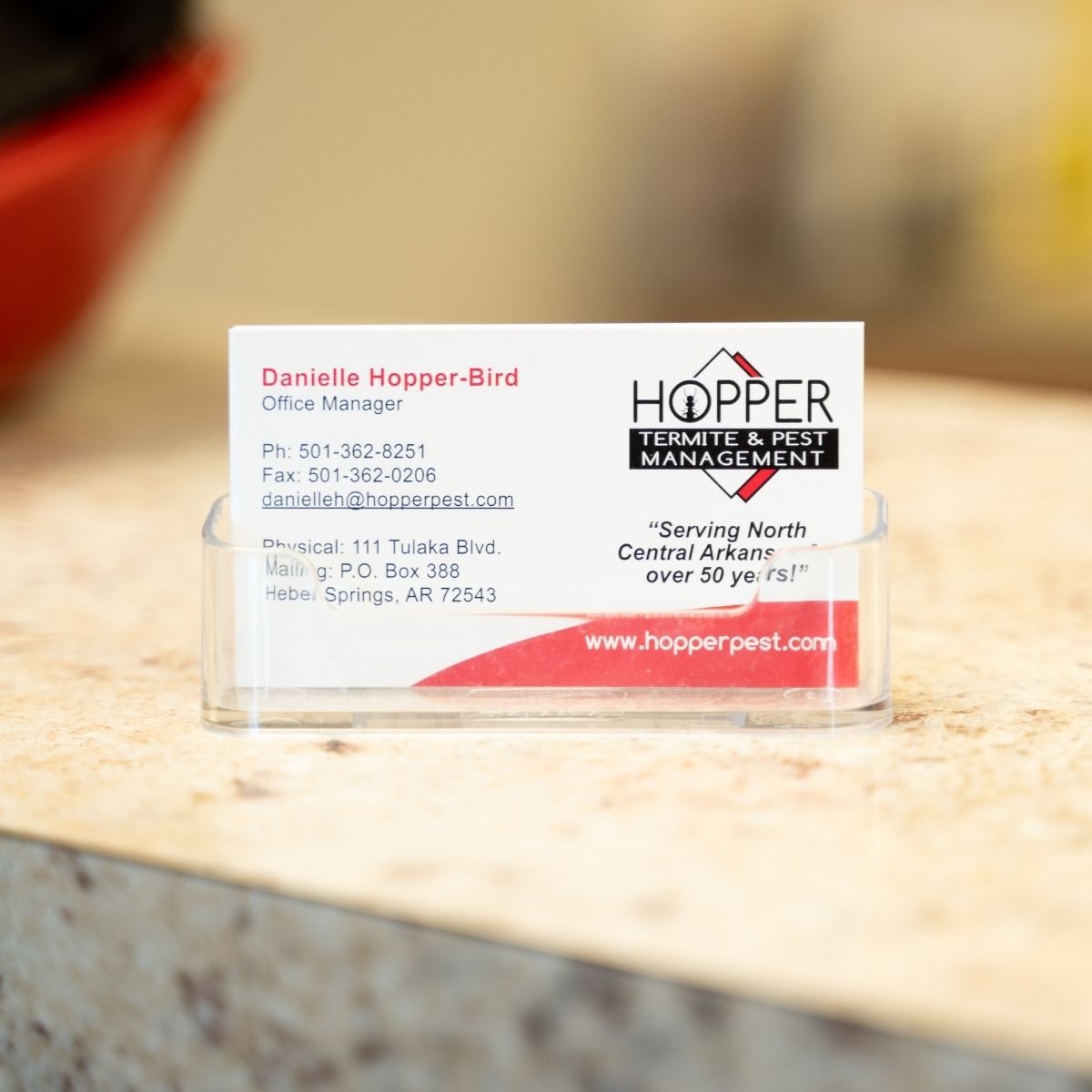 A business card for Danielle Hopper-Bird, Office Manager at Hopper Termite & Pest Management, displayed in a clear holder on a beige countertop. The card includes contact details and the company’s logo.