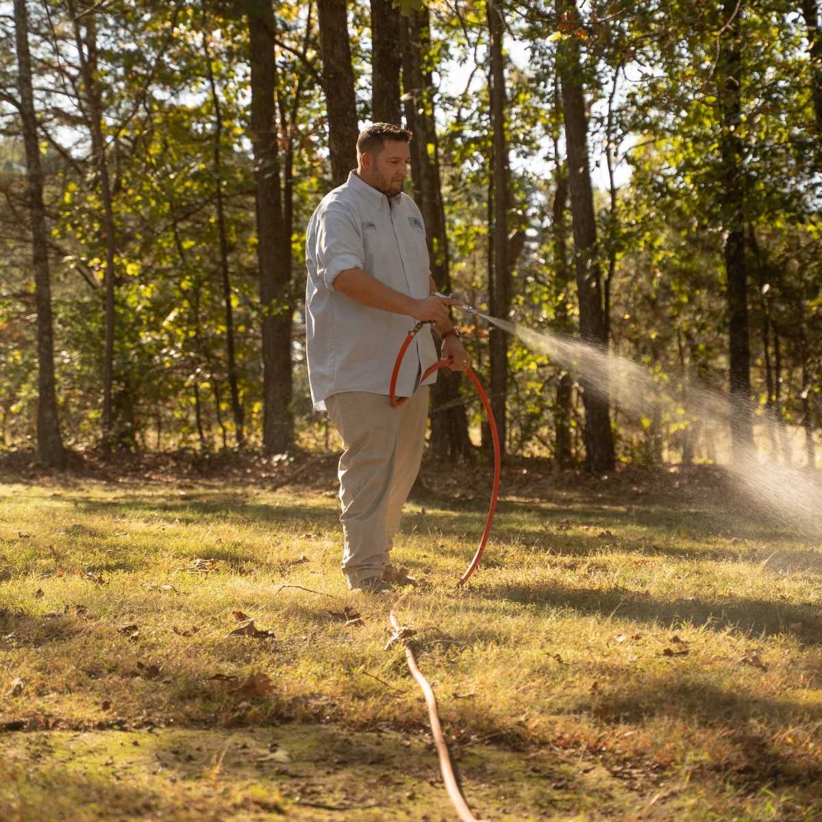 A man stands on a grassy area near trees, holding a red hose and spraying water onto the ground on a sunny day.