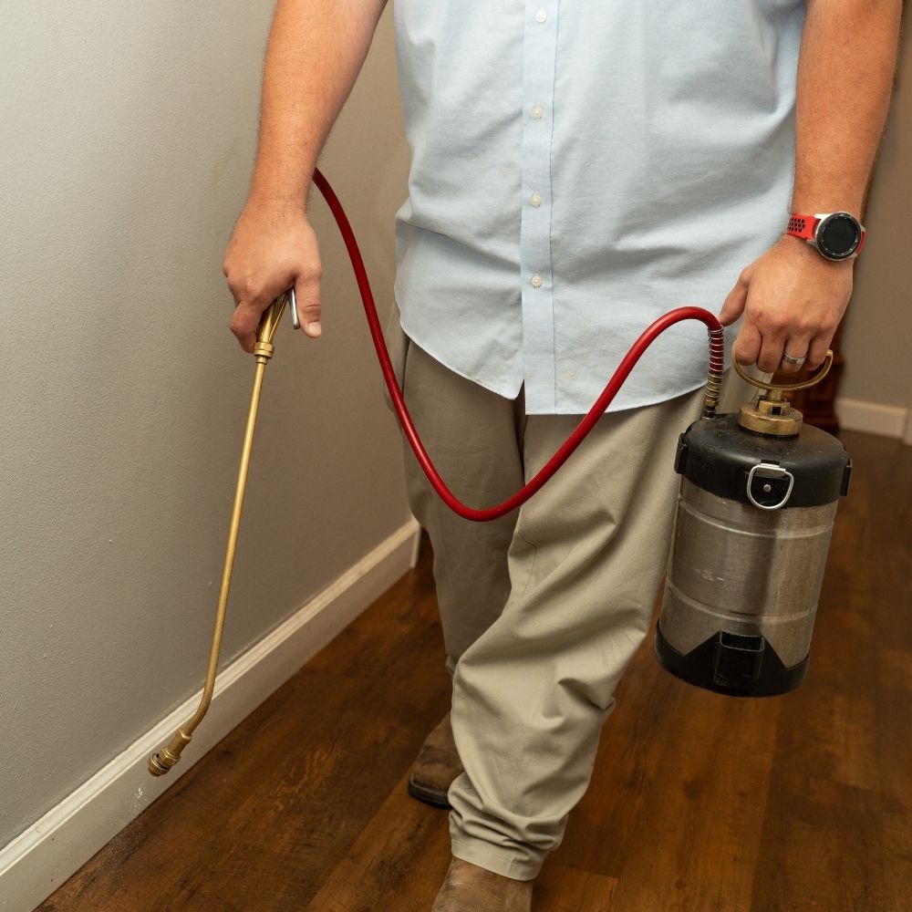 A person in a light blue shirt and khaki pants holds a metal canister sprayer with a red hose, spraying along the baseboard of a room with wood flooring.
