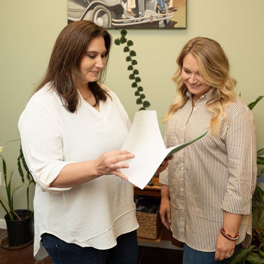 Two women stand together indoors, smiling as one shows the other a document. They are casually dressed, with plants and a framed photo in the background, suggesting a relaxed, friendly environment.
